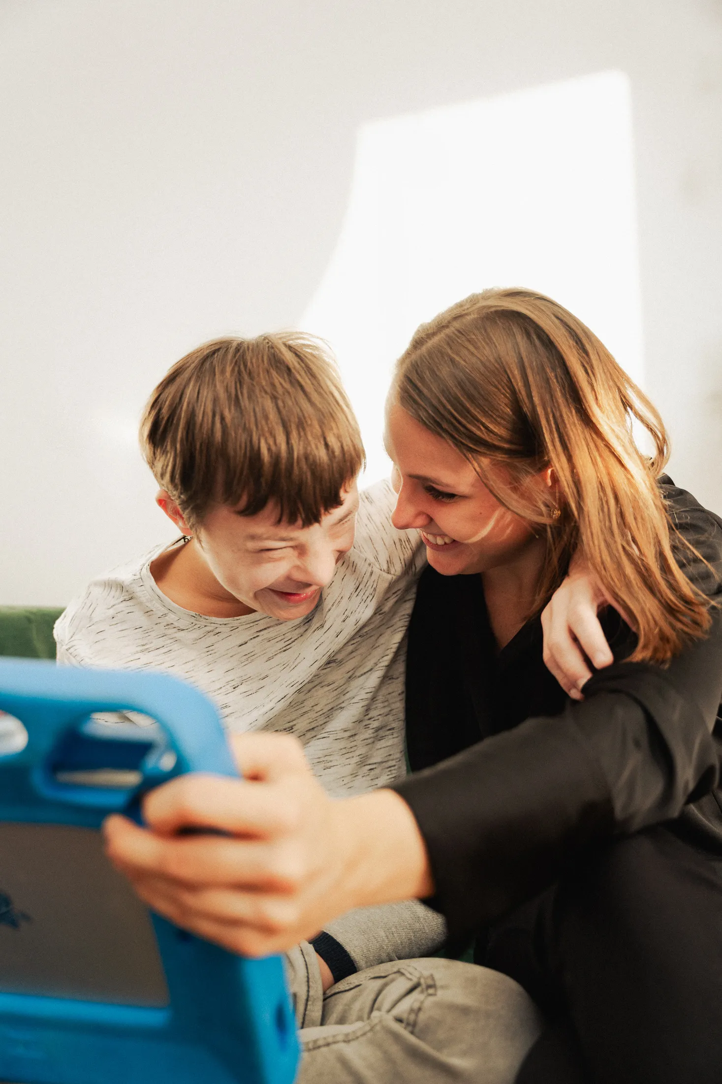 Zwei Personen sitzen nebeneinander und lachen miteinander; im Vordergrund hält eine von ihnen ein Tablet in einer blauen Schutzhülle.