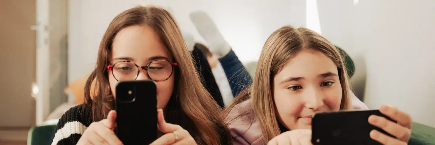 Isabell und Stella liegen nebeneinander auf einer dunkelgrünen Couch auf dem Bauch und schauen auf ihre
Smartphones.